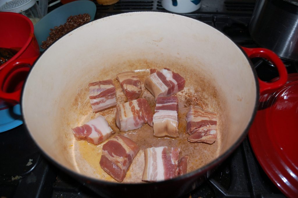 cast iron enameled pan with large chi=unks of bacon being fried.