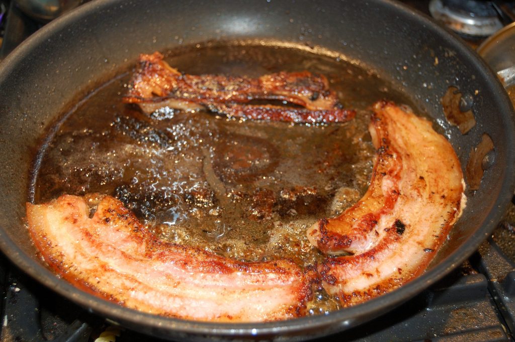 Streaky bacon frying in a pan, nice golden colour to the fat, nice browning to the flesh.